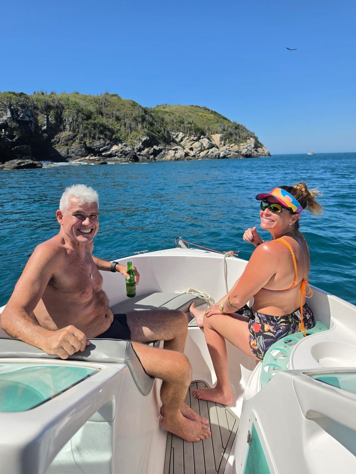 Casal sorrindo na proa da lancha, com uma ilha e o mar azul ao fundo